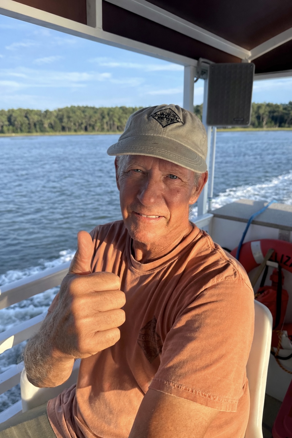 Man on a boat giving a thumbs-up, wearing a cap and orange shirt with water in the background.