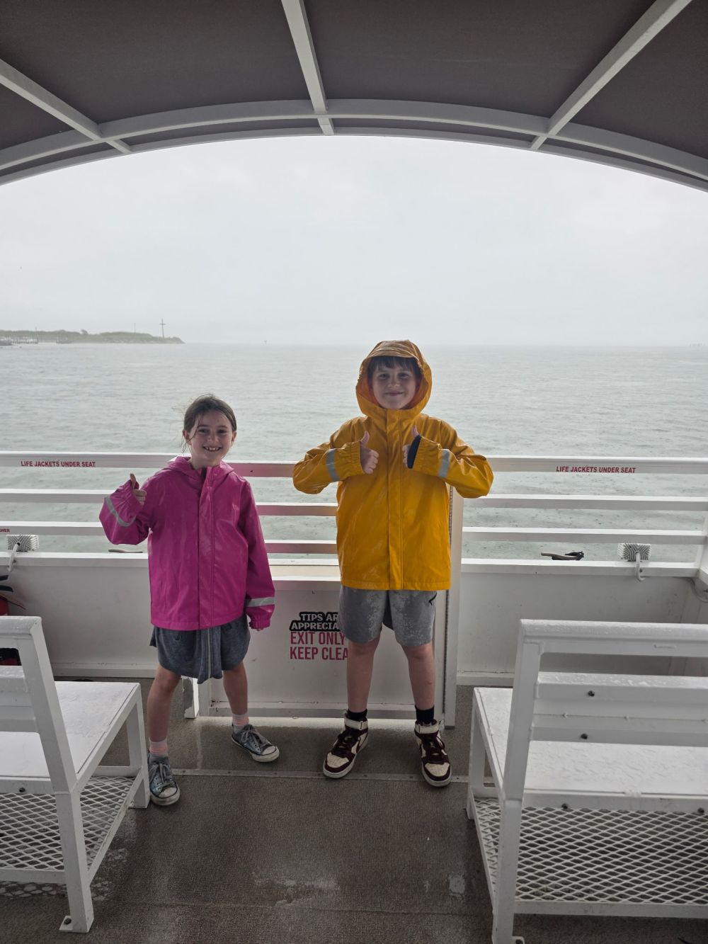 Two kids in raincoats on a ferry with ocean in the background.