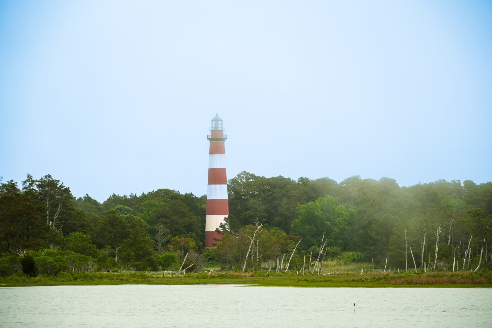 Red and white striped lighthouse near trees with a calm river in foreground.