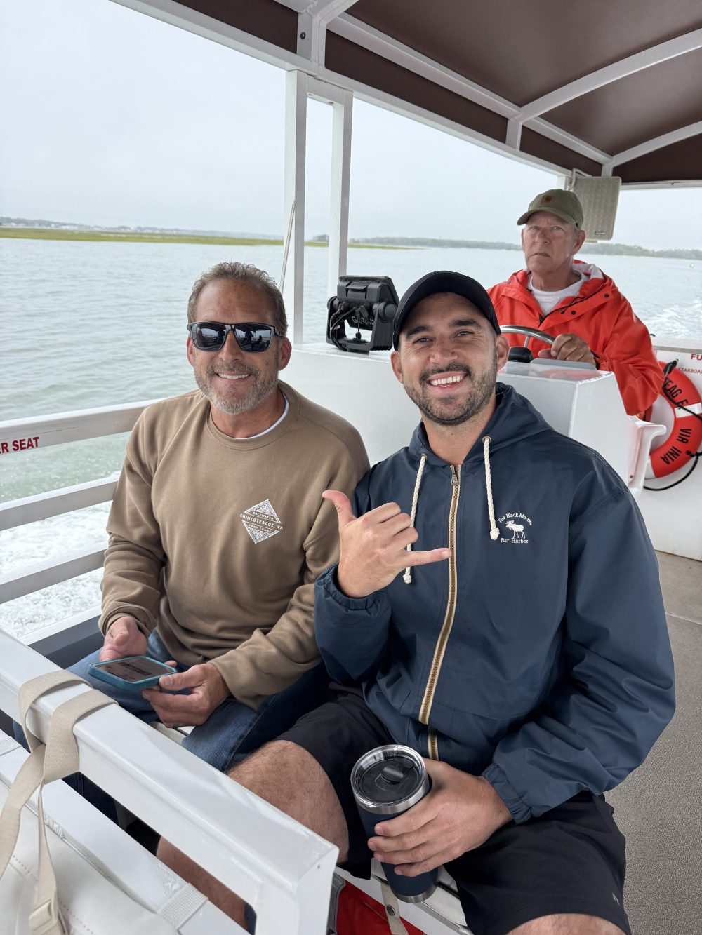 Three men sitting on a boat, one smiling and gesturing, with water in the background.